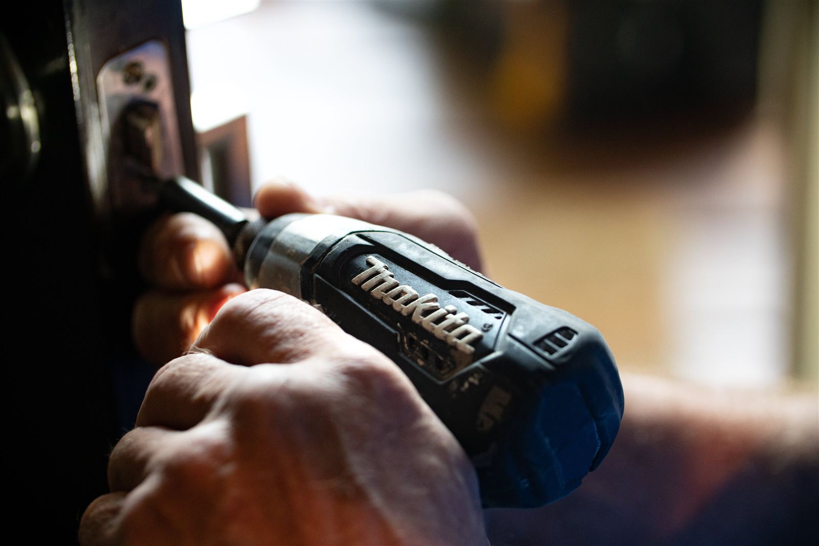 A handyman's hands driving a screw into a door latch with a cordless drill